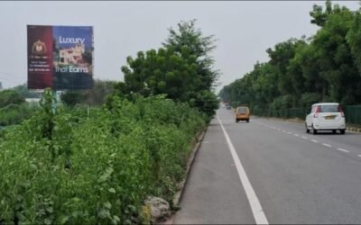 Top Hoarding Advertising Companies in Lucknow | Acme Advertising Co. Highway hoarding in Lucknow placed on a green roadside with moving vehicles, managed by top hoarding advertising companies in Lucknow.