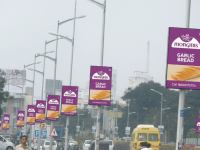 Acme Advertising Co. – Pole Kiosk advertising companies in Lucknow Ads for Bakery Products A row of digital pole kiosk advertising companies in Lucknow displaying advertisements for 'Monginis Garlic Bread,' showcasing images of garlic bread slices, set along a busy highway.